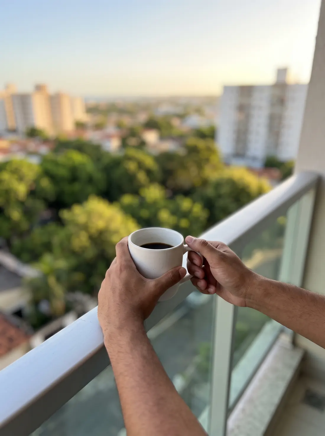 Mãos segurando uma xícara de café na varanda, com a cidade ao fundo